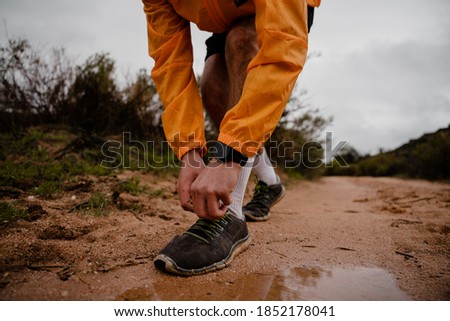 Young fit male athlete bending over tying shoe laces of running shoes while running on wet mountain path in cloudy weather
