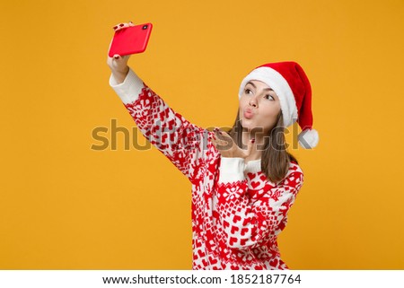 Pretty young Santa woman in sweater Christmas hat doing selfie shot on mobile phone blowing sending air kiss isolated on yellow background, studio portrait. Happy New Year celebration holiday concept