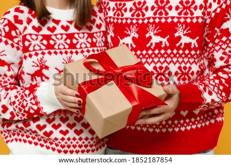 Cropped image of young Santa couple friends man woman in Christmas red sweater hold present box with gift ribbon bow isolated on yellow background studio. Happy New Year celebration merry concept