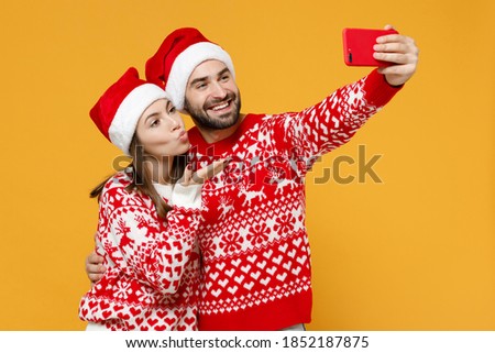 Pretty young Santa couple friends man woman 20s in Christmas hat doing selfie shot on mobile phone blowing sending air kiss isolated on yellow background. Happy New Year celebration holiday concept