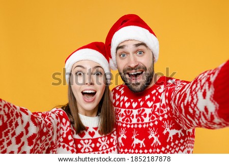 Close up excited young Santa couple friends man woman in red sweater, Christmas hat doing selfie shot on mobile phone isolated on yellow background. Happy New Year celebration merry holiday concept