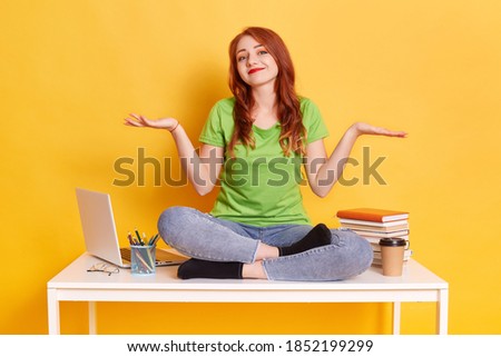 Woman sitting on table near notebook, stack of books, coffee and pens, having break from working on laptop, drinking coffee, spreading hands aside over yellow wall, woman in casual clothing.