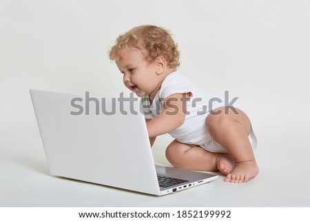 Adorable baby sitting on floor and looking at laptop screen with exited facial expression, infant wearing white body suit, having blond curly hair , touching keyboard with hand.