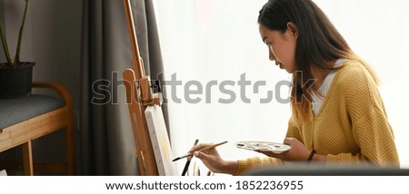 A young female artist in yellow sweater is painting on easel while enjoying work in art studio.