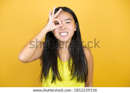 Young beautiful asian woman over yellow isolated background doing ok gesture shocked with smiling face, eye looking through fingers
