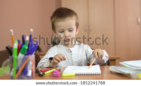 A handsome three-year-old boy sits at a desk in the class and shows off markers and pens. Preschool education. 
