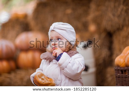 Little girl in beige coat eating croissant on a backgrounds of pumpkins. Childhood concept.