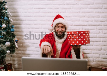 Happy young Santa Claus wearing hat holding gift box using laptop computer sitting at workshop home table on Merry Christmas eve. Ecommerce website xmas time holiday online shopping e commerce sale