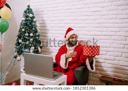 Young Santa Claus with hat video calling kid, talking to child greeting on Merry Christmas, Happy New Year in virtual video online chat on laptop sitting at home table late with present on xmas eve.