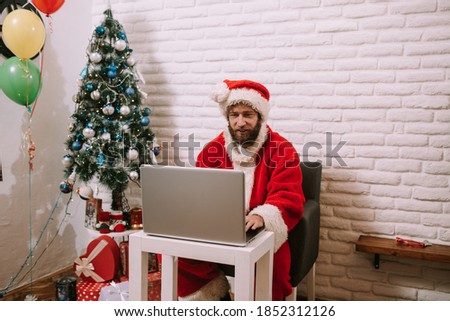 Modern young Santa Claus at his house surfing in the Internet with his laptop. Christmas time. Happy new year
