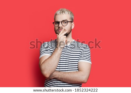 Blonde caucasian man with beard and glasses is posing thoughtful touching his chin on a red studio wall looking at camera