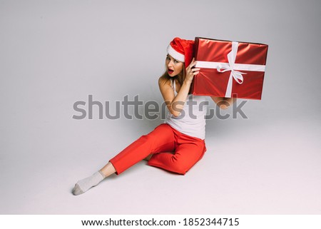 funny woman with christmas hat sits with a big red box and try to hear what is inside, picture isolated on white background