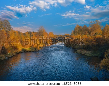 Autumn landscape, forest trees are reflected in calm river water against a background of blue sky and clouds.