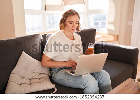 Waist up portrait view of the calm caucasian female sitting at the sofa with her laptop computer while browsing internet. Technology concept. Stock photo