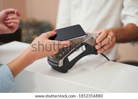 Close up view of the adorable blonde client woman paying with card contactless in the hairdressing salon while standing at the reception. Stock photo