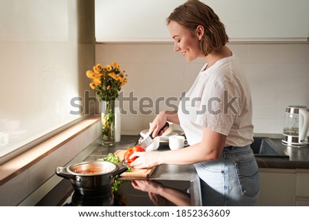Waist up portrait of the pleasant adorable girl preparing vegetables for the future soup while standing at the light kitchen.Stock photo