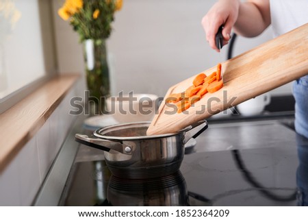 Close up view of the caucasian woman sprinkles carrot from wooden plank to the aluminum saucepan standing on the stove at the kitchen surface. Stock photo
