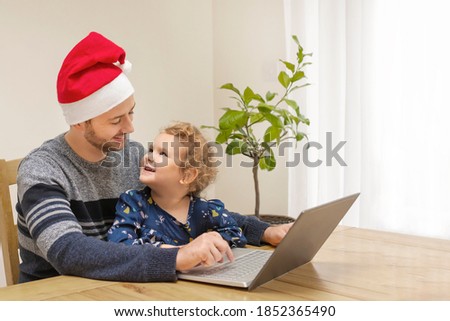 Happy father wearing santa's red hat and little daughter looking at each other laughing using laptop, cheerful dad and child girl having fun playing online game together, watching video, shopping.