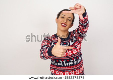 Beautiful arab girl wearing christmas sweater over isolated white background making finger frame with hands. Creativity and photography concept.