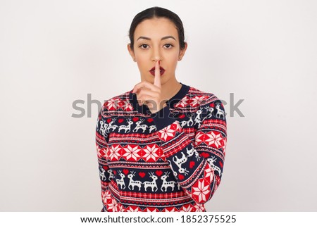 Beautiful arab girl wearing christmas sweater over isolated white background makes silence gesture, keeps finger over lips. Silence and secret concept.