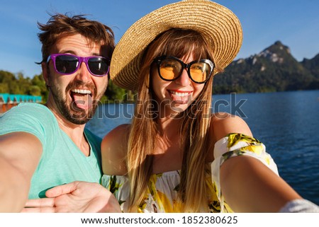 Happy lovely couple making selfie at vacation on mountains and lake, summer bright clothes hat and sunglasses, showing tongues, positive mood.