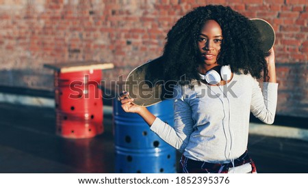 Close up of young beautiful woman with curly hair with white headphones. Hipster girl holding skate over her shoulders and posing for camera outside.