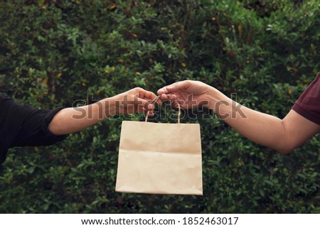 Woman’s hand pick up taking empty craft brown recycled paper shopping bag from man with outdoor green leaves nature hedge background. Delivery paper bag mockup for branding design concept Royalty-Free Stock Photo #1852463017