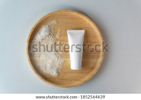 Top down view of organic gentle facial skincare product cleanser white tube with soap bubbles and blank label on bamboo wooden plate grey blue cardboard box paper background. Royalty-Free Stock Photo #1852564639