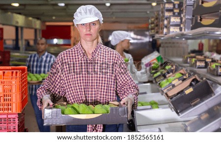 Portrait of positive woman employee working at fruit warehouse carrying box with pears
