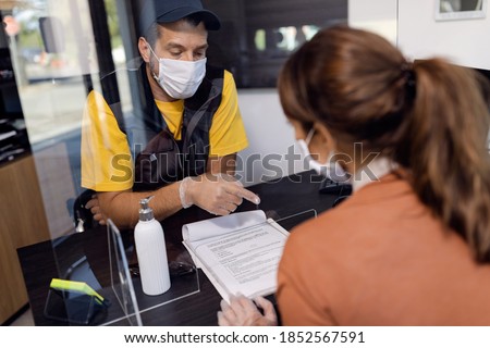 Cam mechanic pointing on place of signature on a document while talking with customer through sneeze guard at workshop's office during coronavirus pandemic. 