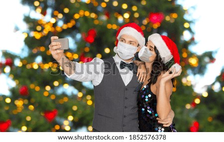 pandemic, health and winter holidays concept - couple wearing face protective medical masks for protection from virus disease taking selfie by smartphone over christmas tree lights on background