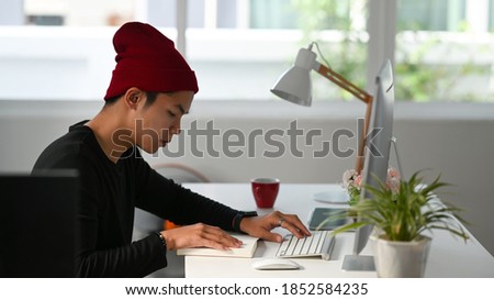Side view of freelance graphic designer sitting at his workspace and looking at notebook while working on computer.