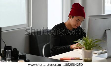 A male creative graphic designer is reading a book while sitting in front of computer at workplace.
