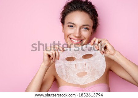 Smiling woman holds facial sheet mask. Photo of attractive woman with perfect skin on pink background. Beauty & Skin care concept Royalty-Free Stock Photo #1852613794