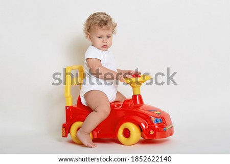 Cute little boy riding on red and yellow toy car, looking away, posing barefoot, dresses bodysuit, has beautiful blond hair, playing with his tolocar isolated over white background.