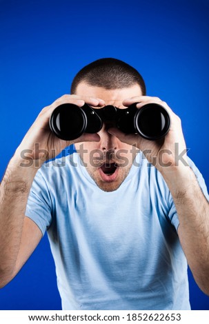 Man with dark short hair looking through binoculars with a surprised look.