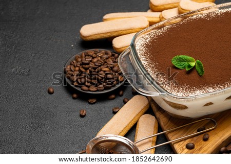 Traditional Italian Tiramisu dessert in glass baking dish on wooden cutting board and savoiardi cookies on concrete background