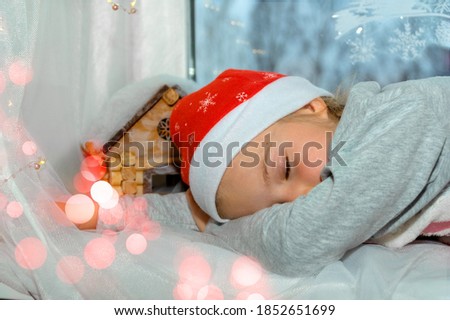 A little girl in a New Year's cap fell asleep on the windowsill. The window is decorated for Christmas. Celebrating Christmas at home.
