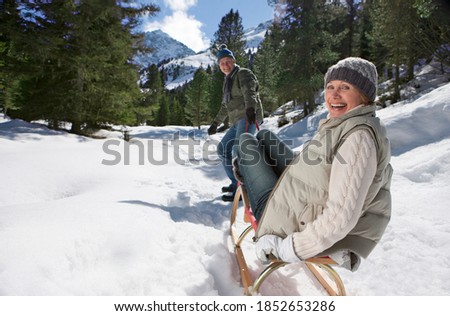 A wide view of a happy woman sitting on a sled and turning back while being pulled by her husband on a snowy slope in the wilderness
