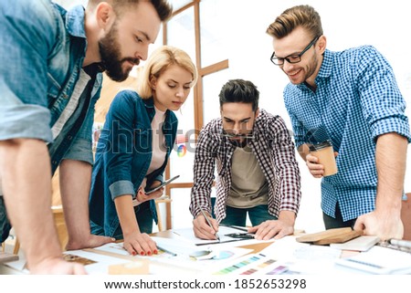 A group of designers stand and draw a new plan for the construction of the complex. Designer in glasses with coffee in hand looks like a colleague is drawing a plan.