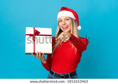 Woman with christmas hat holding a present isolated on blue background points finger at you with a confident expression