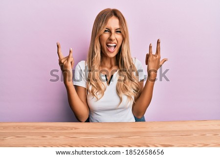 Beautiful blonde young woman wearing casual clothes sitting on the table shouting with crazy expression doing rock symbol with hands up. music star. heavy concept. 