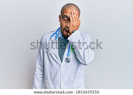 Hispanic adult man wearing doctor uniform and stethoscope yawning tired covering half face, eye and mouth with hand. face hurts in pain. 