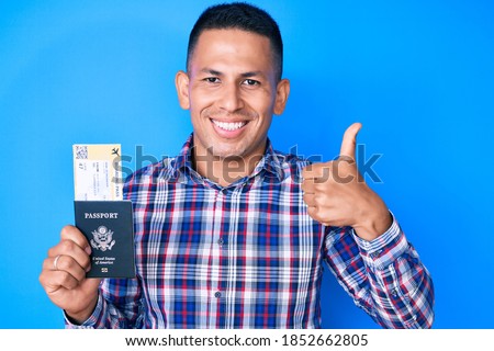 Young handsome latin man holding united states passport and boarding pass smiling happy and positive, thumb up doing excellent and approval sign 