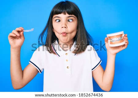 Young little girl with bang holding invisible aligner orthodontic and braces making fish face with mouth and squinting eyes, crazy and comical. 