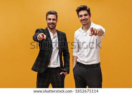 Good-humored young brunet businessmen in white shirts and black pants point at camera abs smile on isolated orange background.