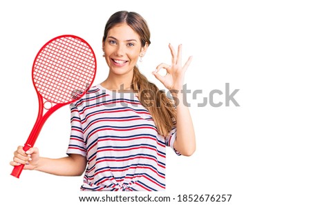 Beautiful caucasian woman with blonde hair playing tennis holding racket doing ok sign with fingers, smiling friendly gesturing excellent symbol 