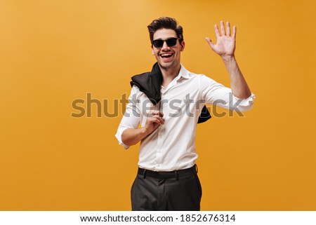 Excited charming brunet man in white shirt, sunglasses and black pants smiles, holds jacket and waves hand in greeting on orange background.
