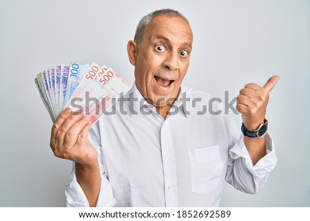 Handsome senior man holding swedish krona banknotes pointing thumb up to the side smiling happy with open mouth  Royalty-Free Stock Photo #1852692589