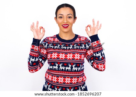 Glad Young beautiful Arab woman wearing Christmas sweater against white wall shows ok sign with both hands as expresses approval, has cheerful expression, being optimistic.